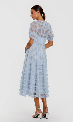 A woman stands facing slightly away from the camera in the Mac Duggal 8288 Midi Party Dress, a light blue a-line midi dress with sheer short sleeves and layered ruffles. She pairs it with silver block-heeled sandals and wears her hair in a low ponytail.