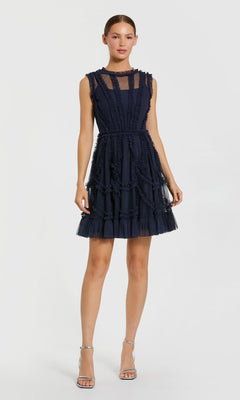 A woman stands against a white background wearing the Mac Duggal 8289 short wedding guest dress in indigo, a sleeveless fit and flare style with sheer paneling and ruffle details, paired with silver high-heeled sandals.