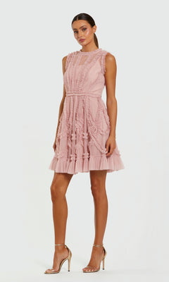 A woman stands against a plain white background in a rose Mac Duggal 8289 short wedding guest dress with ruffle details and strappy high-heeled sandals.
