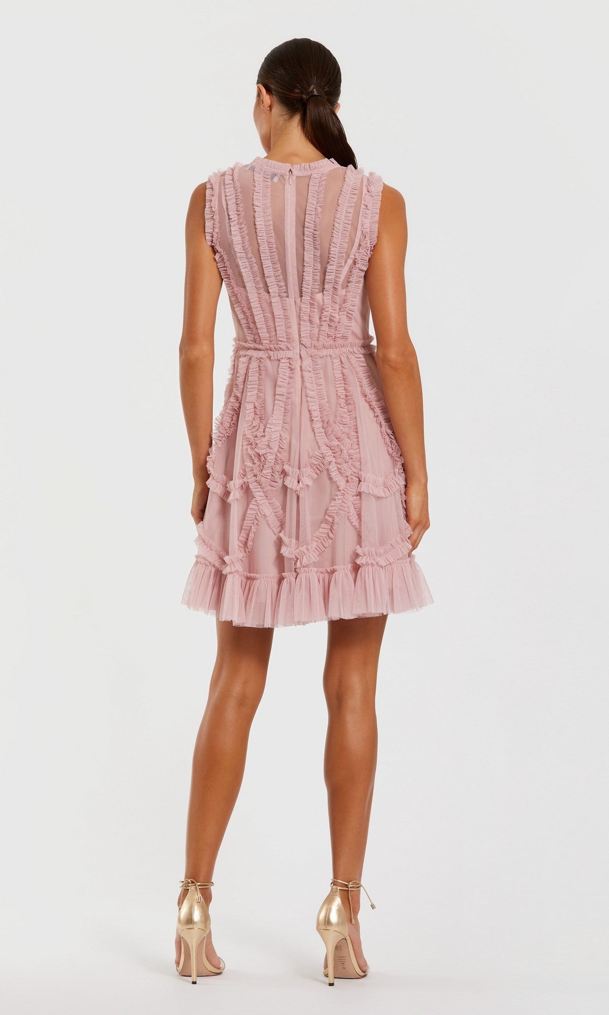 Woman in the Mac Duggal 8289 Short Wedding Guest Dress, a sleeveless high-neck mini with pink ruffles and sheer panels, stands with her back to the camera wearing gold high-heeled shoes.