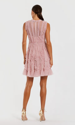 Woman in the Mac Duggal 8289 Short Wedding Guest Dress, a sleeveless high-neck mini with pink ruffles and sheer panels, stands with her back to the camera wearing gold high-heeled shoes.