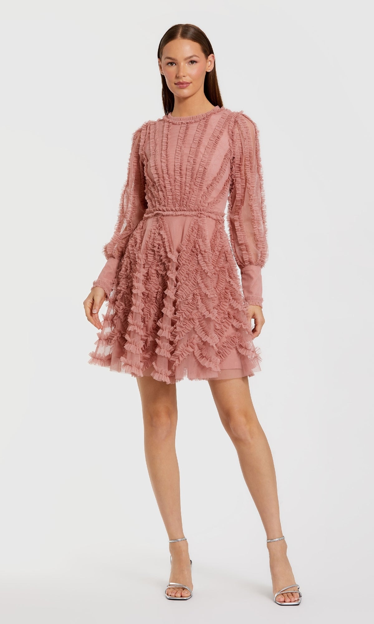 Woman wearing a short pink dress with long sleeves and mini ruffles, standing in front of a white background