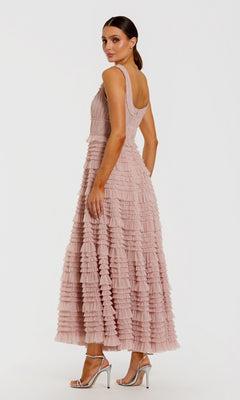 A woman stands in the Tea-Length Party Dress: Mac Duggal 8292—a sleeveless, blush pink mesh midi cocktail dress featuring delicate ruffle detailing and an elegant A-line tiered skirt—paired with silver high-heeled sandals, facing slightly away against a plain white background.