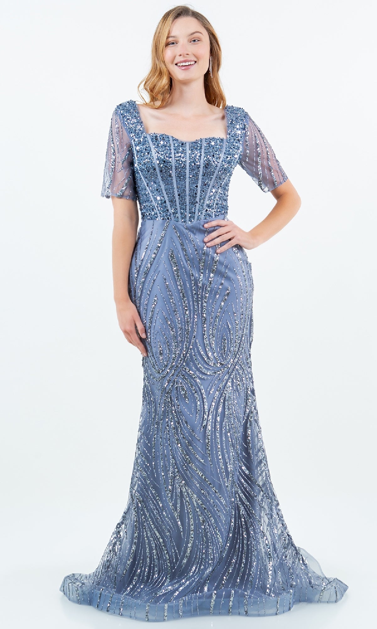 A woman models the Cinderella Couture 8293M, a blue-gray sequin formal gown with sheer short sleeves, a square neckline, and bead embellishments. The floor-length dress offers an elegant evening look against a plain white background.