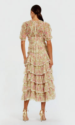 A woman stands facing away, wearing the Midi Wedding Guest Dress: Mac Duggal 8295, a pink and cream floral mesh midi dress with ruffle details, puff sleeves, and gold high-heeled sandals.