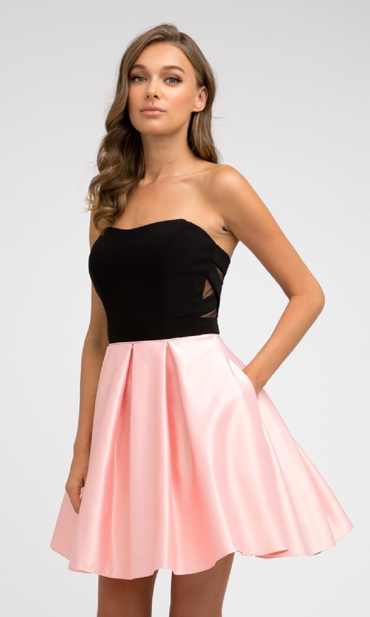 A woman wears the Juliet 829 Short Party Dress, featuring a strapless sweetheart neckline, black fitted bodice, and light pink A-line skirt, standing with one hand in her pocket against a plain background.