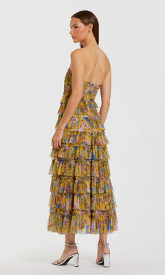 A woman faces sideways wearing the Mac Duggal 8301 Yellow Floral Wedding-Guest Dress, a strapless midi with tiered ruffles, colorful abstract patterns, and silver high-heeled sandals.