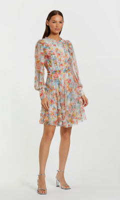 A woman stands against a light background wearing the Short Wedding Guest Dress: Mac Duggal 8304 in Blue Multi—a long-sleeve, floral mesh dress with a fitted waist—paired with silver high-heeled sandals.