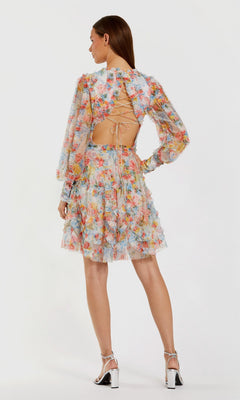 Woman models the Mac Duggal 8304 short wedding guest dress featuring a floral mesh design, long sleeves, and an open back with crisscross laces, paired with high-heeled sandals and facing away from the camera.