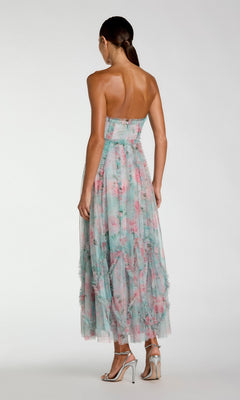 A woman faces away, wearing the Mac Duggal 8308 Strapless Print Midi Party Dress in pastel florals and silver high-heeled sandals, standing against a plain background.