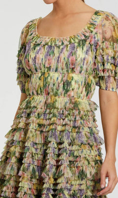 A person wearing the Mac Duggal 8327 Short Party Dress, featuring a square neckline, short sleeves, layered ruffles, and a mini length with a multicolored abstract floral print.