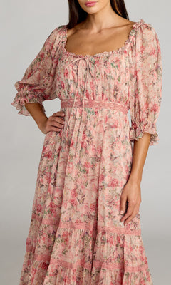 A woman poses with one hand on her hip, wearing the Mac Duggal 8330—Tea-Length Party Dress—a long, pink floral party dress featuring puffed three-quarter sleeves and a square neckline.