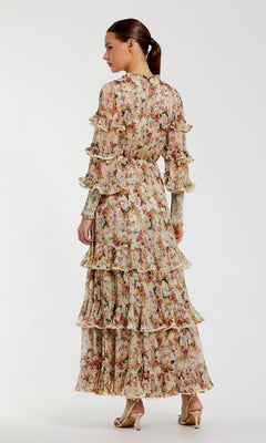 A woman stands facing away in the Mac Duggal 8334 Long Sleeve Ruffle Formal Dress, featuring a floral print, sheer sleeves, and tiered skirt; her hair is tied back and she's wearing heeled sandals against a plain background.