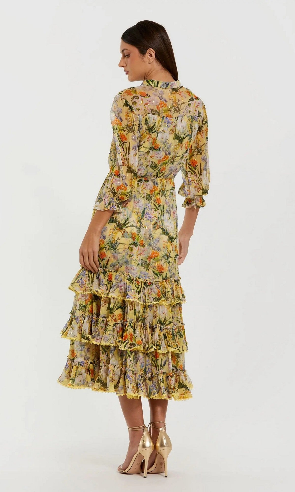 A woman models the Mac Duggal 8335 Midi Party Dress, showcasing its yellow floral print, long sleeves, and modest midi silhouette with tiered ruffles; she is paired with nude high-heeled sandals and stands facing away against a plain white background.