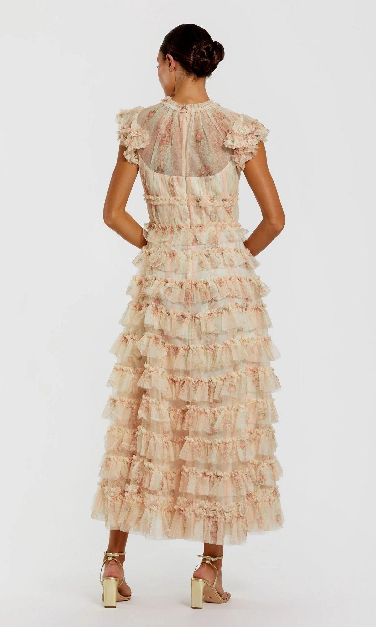 A woman stands facing away from the camera in the Mac Duggal 8339 Tea-Length Party Dress, a sheer, ruffled floral print gown featuring cap sleeves and tiered layers; her hair is styled in a bun and she wears strappy heels.