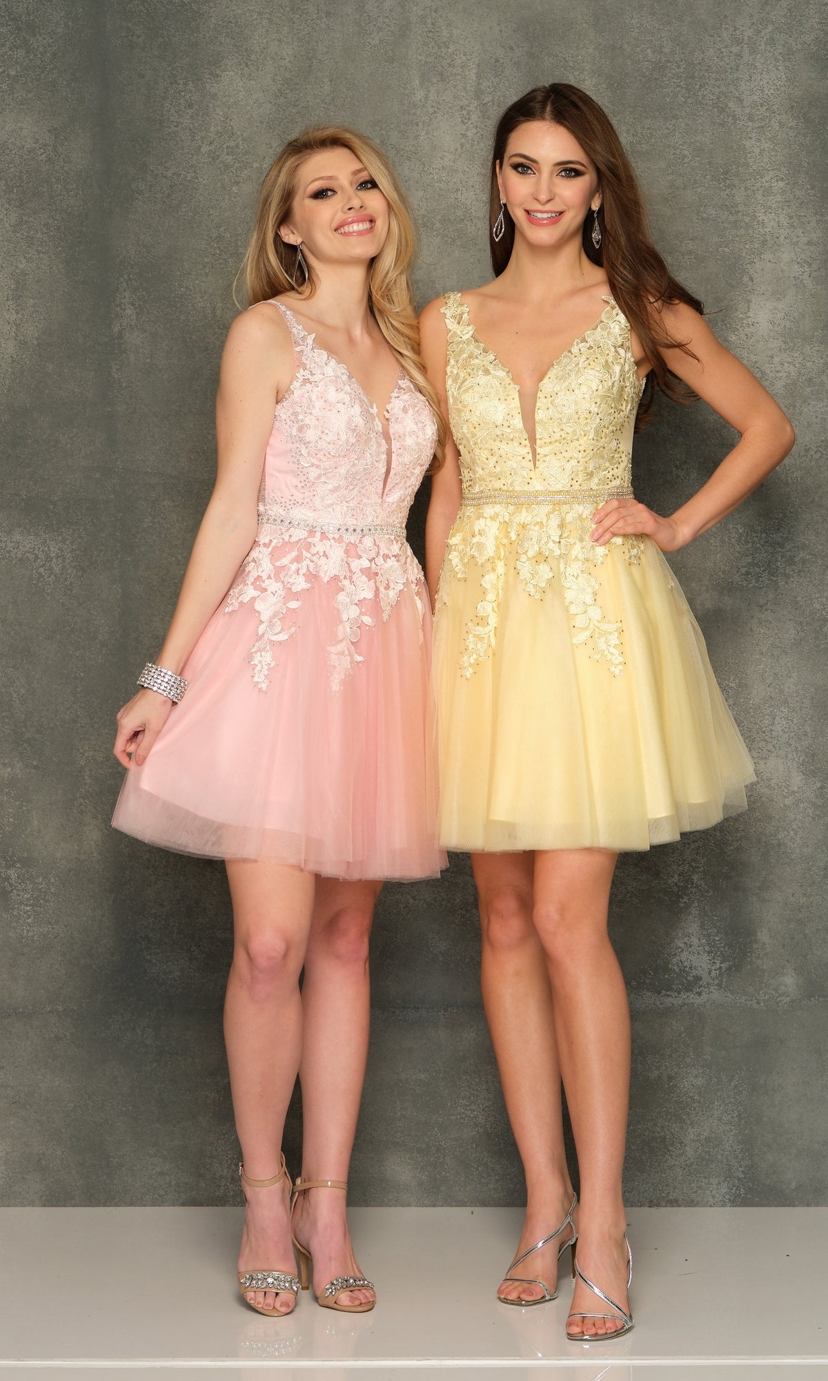 Two women pose before a gray backdrop in A-Line Short Homecoming Dress H341, one pink and one yellow. Each dress features lace detailing and beaded bodices. Both accessorize with heels and earrings for a stylish look.