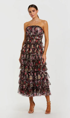 A woman wearing the Mac Duggal 8351 Tea-Length Party Dress in Black Multi—a strapless style featuring a tiered, ruffled silhouette and vibrant floral print—stands against a plain white background, paired with strappy high-heeled sandals.