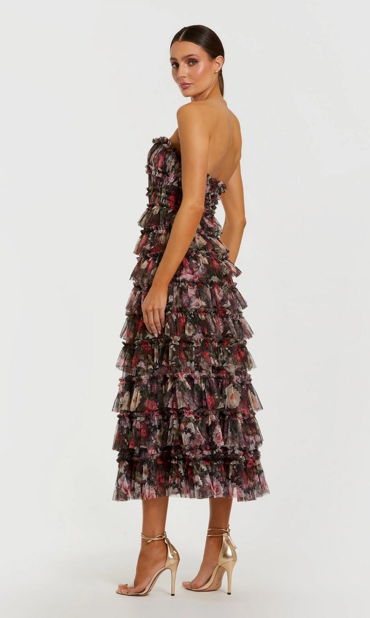A woman poses in the Tea-Length Party Dress: Mac Duggal 8351, a strapless midi dress with tiered ruffle layers and a delicate floral print, paired with gold high-heeled sandals. She stands slightly turned from the camera against a plain light background.