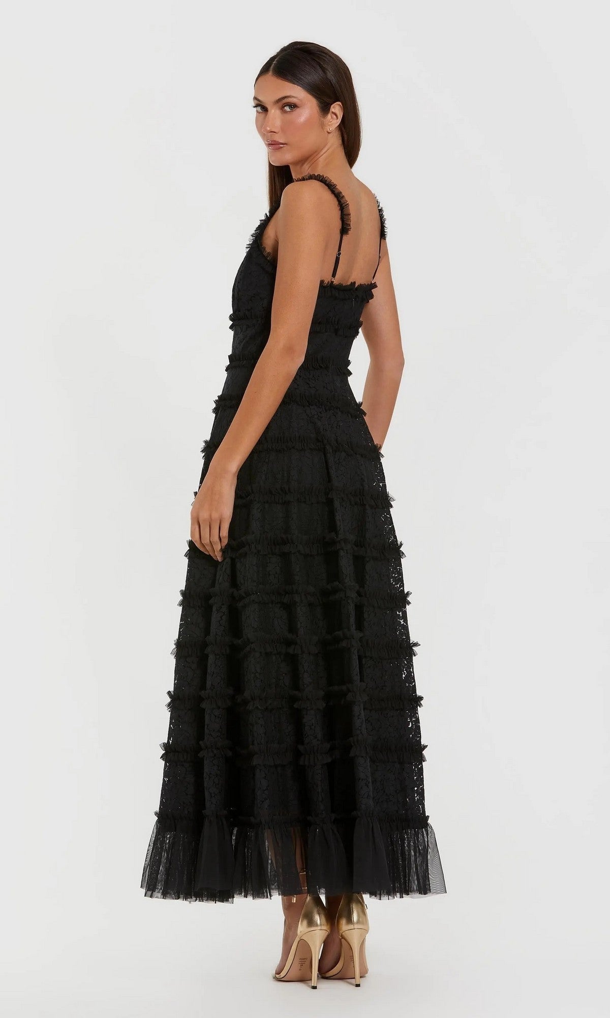 A woman in the Mac Duggal 8372 Floral Lace Long Formal Dress—a sleeveless black lace gown with sheer layers and an ankle-length A-line skirt—stands in heels, facing away and glancing over her shoulder against a white background.