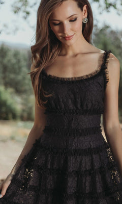 A woman in the Mac Duggal 8372 Floral Lace Long Formal Dress, featuring a sleeveless black floral lace design with ruffle details and a scoop neckline, stands outdoors looking down with a gentle expression.