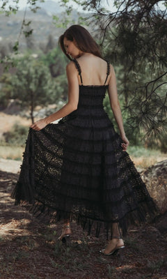 A woman stands outdoors, facing away, holding the sides of her Mac Duggal 8372 Floral Lace Long Formal Dress with a scoop neckline and ankle-length A-line skirt amid trees and natural scenery.