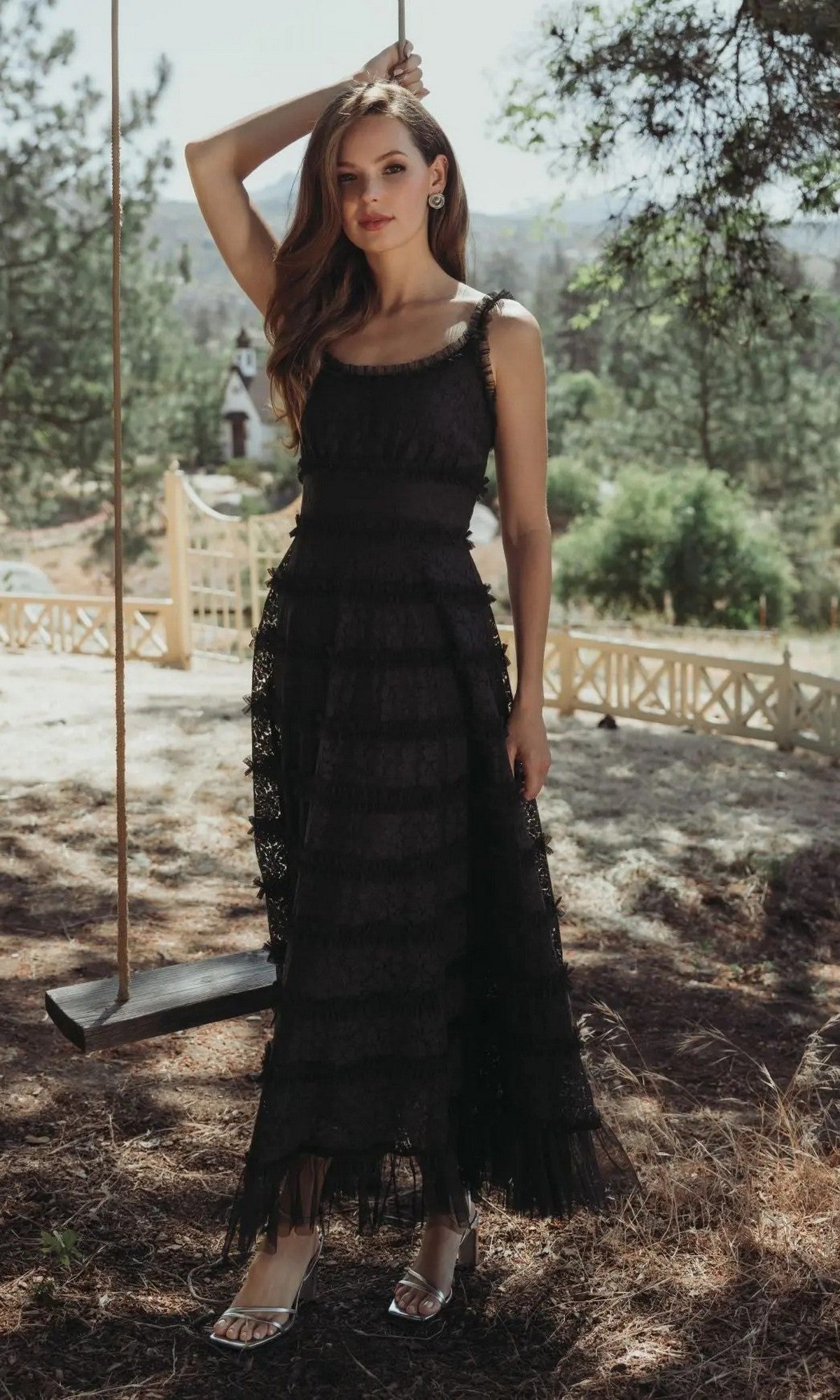 A woman in the Mac Duggal 8372 Floral Lace Long Formal Dress, black, with a sleeveless scoop neckline, stands outdoors by a wooden swing with trees and a fenced yard in the background.