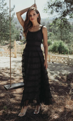 A woman in the Mac Duggal 8372 Floral Lace Long Formal Dress, black, with a sleeveless scoop neckline, stands outdoors by a wooden swing with trees and a fenced yard in the background.