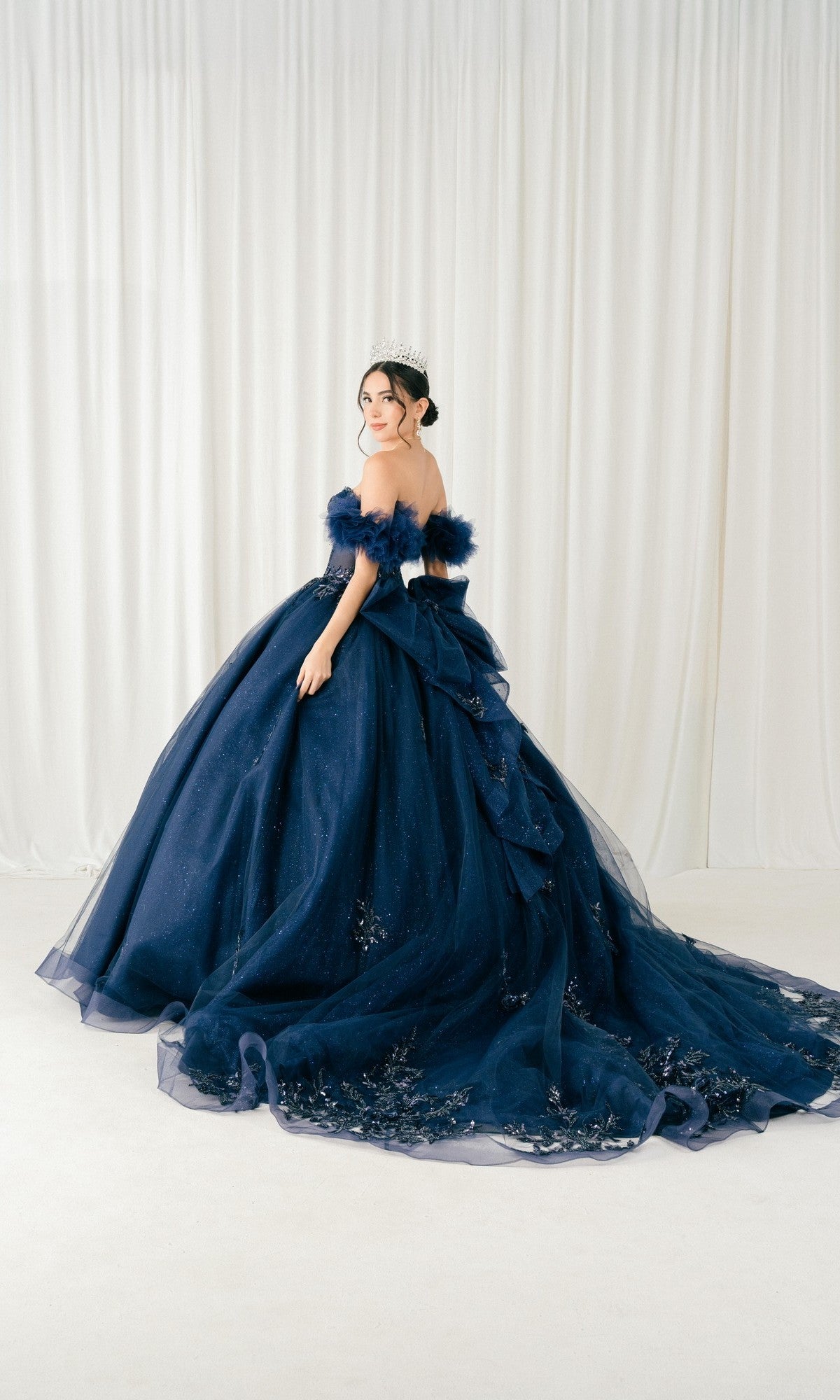 A person wearing the Quinceanera Dress 8380J By Cinderella Couture, a navy blue ball gown with a corset bodice and tiara, stands in front of white curtains with their back turned, looking over their shoulder.