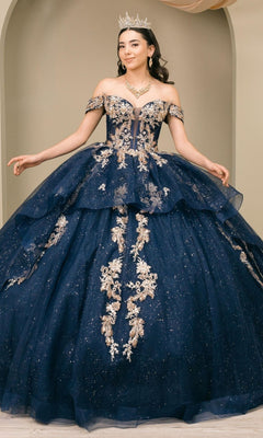 A woman poses elegantly against a beige background, wearing the Quinceanera Dress 8390JQ by Cinderella Couture in navy, adorned with intricate gold embroidery and complemented by a sparkling tiara.