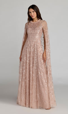 A woman stands against a plain background wearing the Mac Duggal 8415, a rose-colored long formal dress featuring sequins, sheer embellished fabric, and cape-style long sleeves.