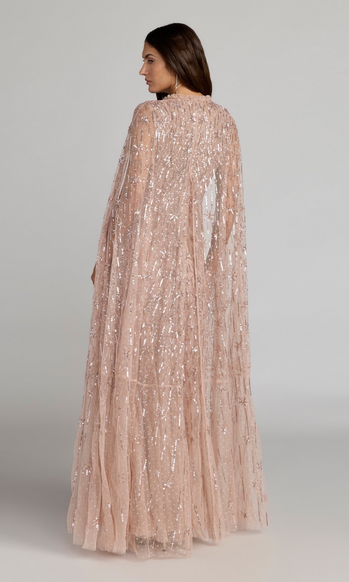 A woman faces sideways in the Mac Duggal 8415 long formal dress, a blush pink sequin evening gown with a sheer, cape-like overlay, set against a plain background.