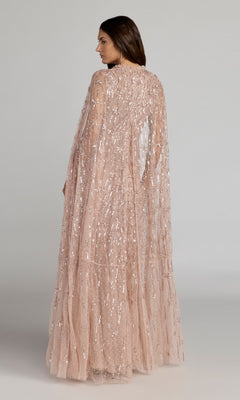 A woman faces sideways in the Mac Duggal 8415 long formal dress, a blush pink sequin evening gown with a sheer, cape-like overlay, set against a plain background.
