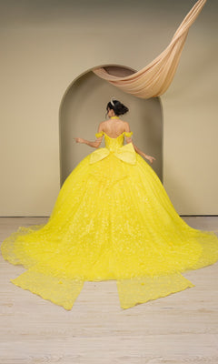 A woman wearing the Quinceanera Dress 8415JQ By Cinderella Couture, an elaborate yellow off-the-shoulder gown, stands with her back to the camera before an arched doorway on a light wooden floor.