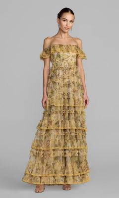 A woman wearing the Mac Duggal 8418 long formal dress in buttercream, featuring floor-length tiers and delicate ruffles with an off-the-shoulder design, stands against a plain gray background.