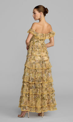 A woman stands in profile against a plain gray background, modeling the Long Formal Dress: Mac Duggal 8418—a yellow floral off-the-shoulder gown with tiered ruffles and a formal A-line silhouette—paired with beige high-heeled sandals.