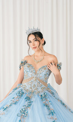 A woman stands before white curtains in the Quinceanera Dress 8420JQ By Cinderella Couture, an ornate light blue ball gown with floral details, sequin and beaded applique, corset back. She wears a tiara, necklace, and her hair is styled up.