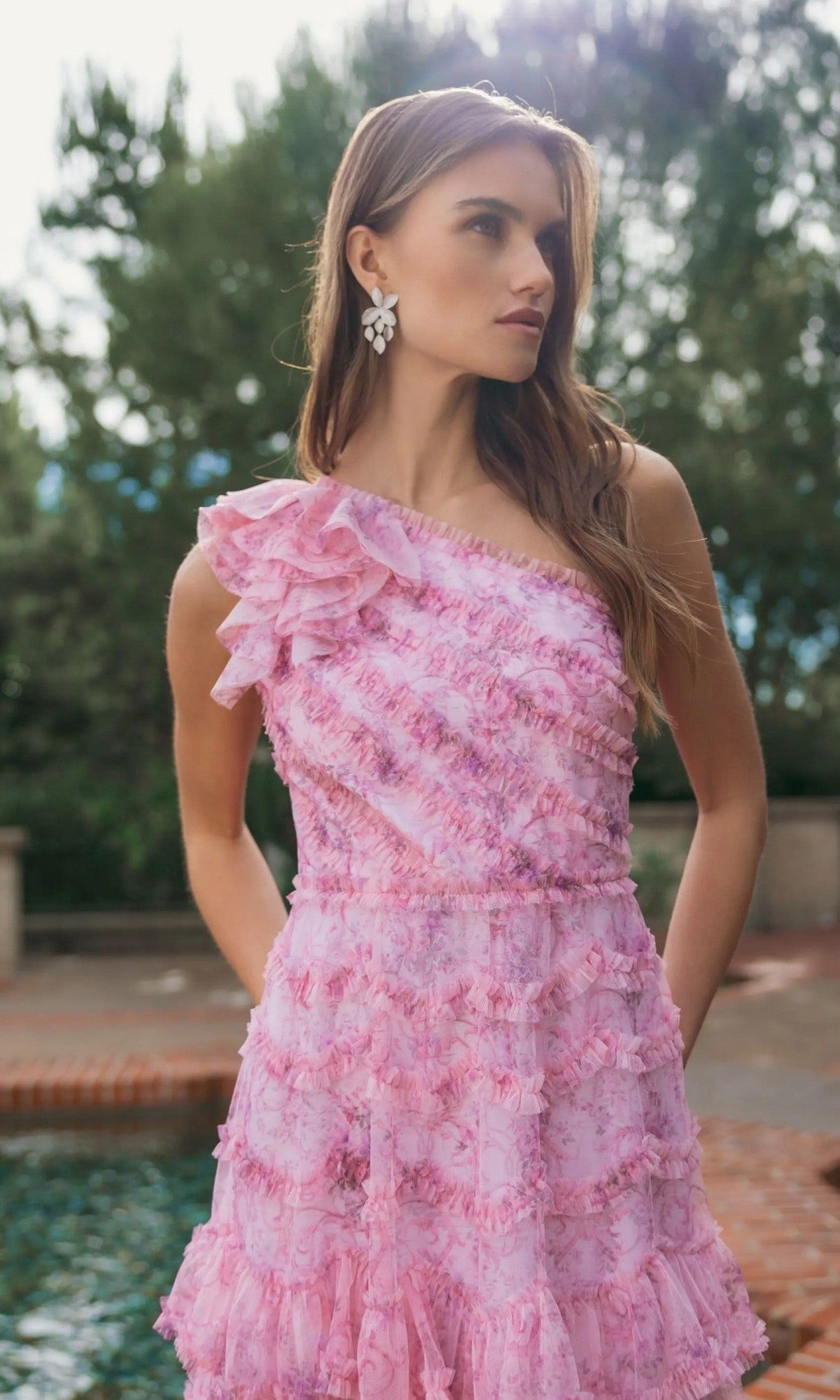 A woman stands outdoors wearing the Mac Duggal 8422 Tea-Length Party Dress—a pink, ruffled, one-shoulder design—paired with white earrings. Trees and a brick-edged fountain in the background complement her elegant ensemble.