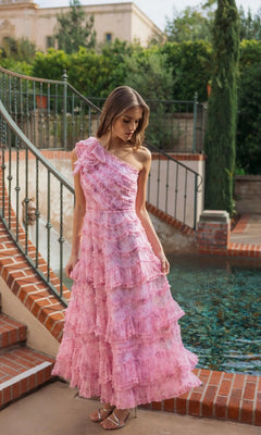 A woman in a pink, ruffled, Mac Duggal 8422 Tea-Length Party Dress stands by a brick-bordered pool with iron railings and greenery in the background.