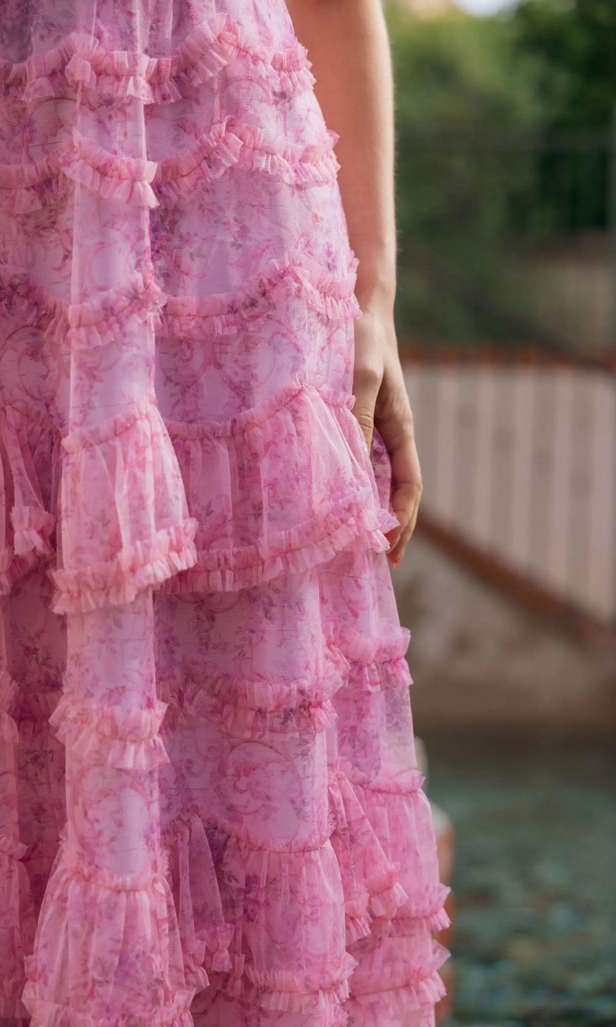 A person stands outdoors wearing the Mac Duggal 8422 Tea-Length Party Dress, featuring pink, ruffled, semi-sheer floral print; only their lower half and one hand are visible.