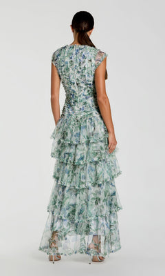 A woman stands facing away, wearing the Long Formal Dress: Mac Duggal 8423—a sleeveless, floor-length A-line gown featuring ruffles and a blue and green floral pattern—paired with high heels against a plain white background.