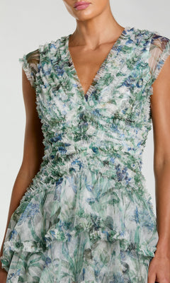 A woman wearing the Mac Duggal 8423 Long Formal Dress, featuring a sleeveless V-neck design with a blue floral pattern in green tones and ruffled detailing throughout the bodice and skirt.