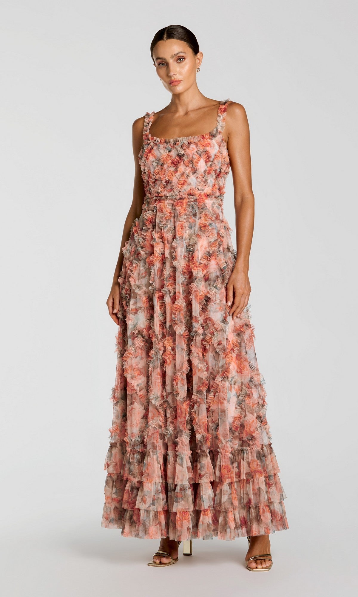 A woman wearing the Mac Duggal 8424 Long Formal Dress in Cream Multi, a sleeveless, floor-length gown featuring ruffle layers and a striking floral pattern, stands against a plain light background.