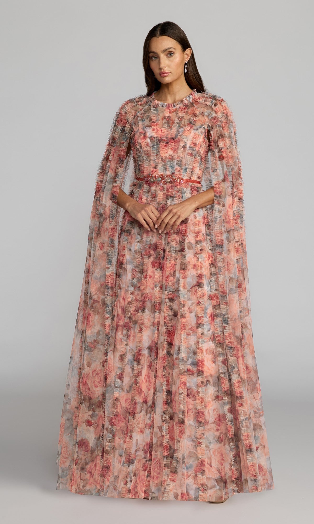A woman wearing the Mac Duggal 8431 long formal dress in cream multi, featuring sheer cape sleeves and pink and red rose floral patterns, stands against a plain gray background.