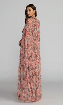 A woman stands looking over her shoulder, wearing the Mac Duggal 8431 long formal dress—a flowy, pink floral gown crafted from sheer fabric—set against a neutral background.