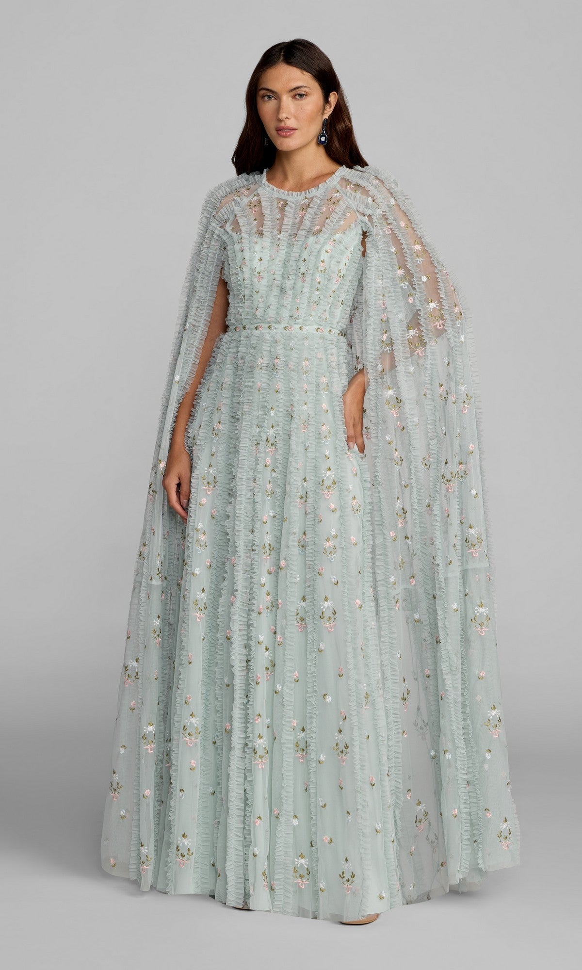 A woman stands wearing the Mac Duggal 8444 Long Formal Dress in a seafoam multi hue, featuring floor-length ruffled details and delicate floral embroidery. A matching sheer cape adds elegance to the floral-embroidered dress.