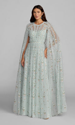 A woman stands wearing the Mac Duggal 8444 Long Formal Dress in a seafoam multi hue, featuring floor-length ruffled details and delicate floral embroidery. A matching sheer cape adds elegance to the floral-embroidered dress.