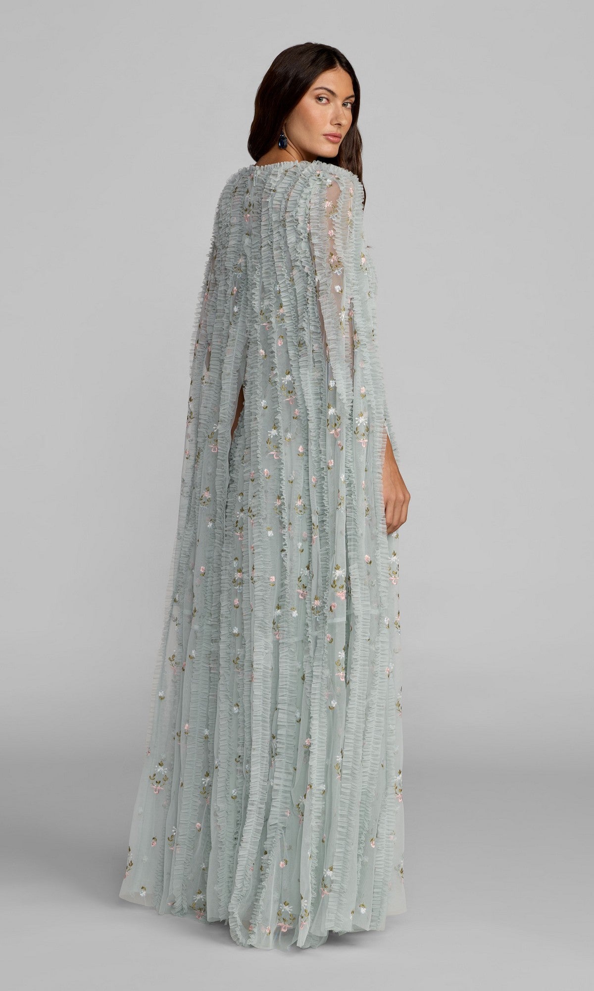 A woman stands on a gray background wearing the Mac Duggal 8444 long formal dress, featuring floor-length light blue floral embroidery, delicate sheer fabric, textured pleats, and intricate detailing as she looks over her shoulder.
