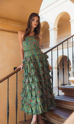 A woman in the green multi Tea-Length Party Dress: Mac Duggal 8459 stands on a staircase, holding the railing, with an arched building in the background.