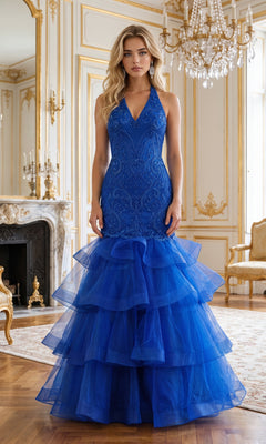 A woman wearing the Beaded Long Halter Mermaid Tiered Prom Dress 8466 in royal stands in an ornate room adorned with gold accents, chandeliers, a marble fireplace, and elegant furniture.