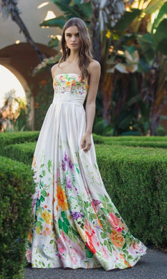 A woman stands outdoors among green hedges and tropical plants, wearing an elegant Mac Duggal 8470 long formal dress in ivory multi, her sleeveless floral gown evoking timeless sophistication.